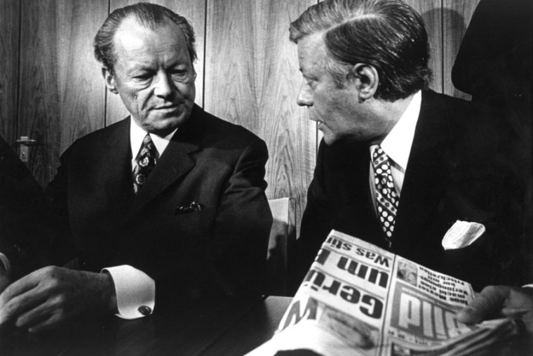 Schwarz-Weiß-Aufnahme von Willy Brandt und Helmut Schmidt. Beide blicken sich an, Schmidt hat eine Ausgabe der Bildzeitung in der Hand.
