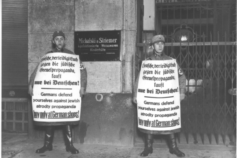 chwarz-Weiß-Aufnahme von zwei SA-Mitgliedern, die vor einem Geschäft stehen und Plakate halten mit „Deutsche, verteidigt euch gegen die jüdische Greuelpropaganda und kauft nur bei Deutschen! Germans defend yourselves against jewish atrocity propaganda buy only at German shops!“.