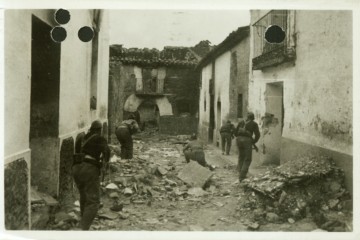 Schwarz-Weiß-Postkarte, auf der fünf Männer mit Gewehren in der Hand zu sehen sind. Sie nähern sich einem verstörten Haus. Auf der Straße neben Ihnen liegt Schutt.