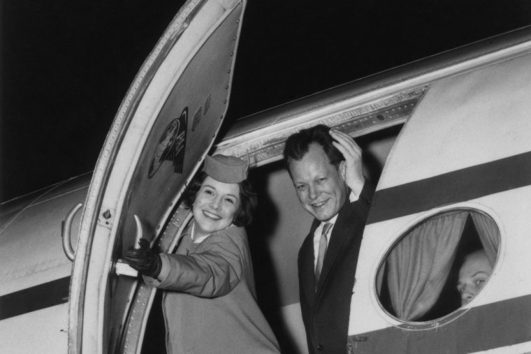 Willy Brandt winkt aus der Tür eines Flugzeuges, während eine Flugbegleiterin diese im Begriff ist zu schließen. Fotografie in Schwarz-Weiß.