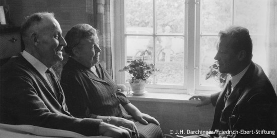 Willy Brandt sitzt bei einem Besuch in Lübeck seiner Mutter Martha Frahm und seinem Stiefvater Emil Kuhlmann gegenüber. Fotografie in Schwarz-Weiß. Öffnet weitere Informationen zu seiner Lübecker Familie.