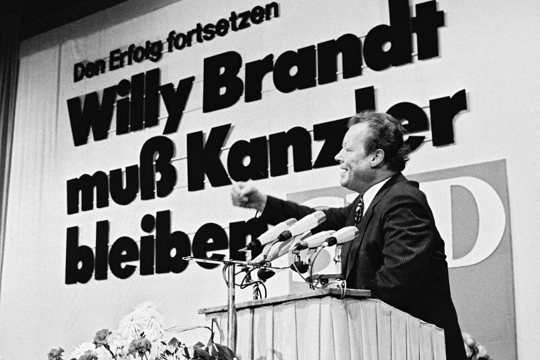 Schwarz-Weiß-Aufnahme Willy Brandts aus dem Jahre 1972 vor einem Rednerpult. Hinter Ihm eine Leinwand auf der steht: „Den Erfolg fortsetzen. Willy Brandt muss Kanzler bleiben.“