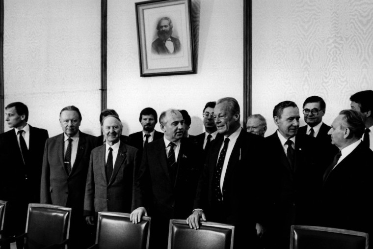 Gruppenbild von Willy Brandt und einer SPD-Delegation beim sowjetischen Generalsekretär Michail Gorbatschow. Im Hintergrund hängt ein Bild von Karl Marx. Fotografie in Schwarz-Weiß.