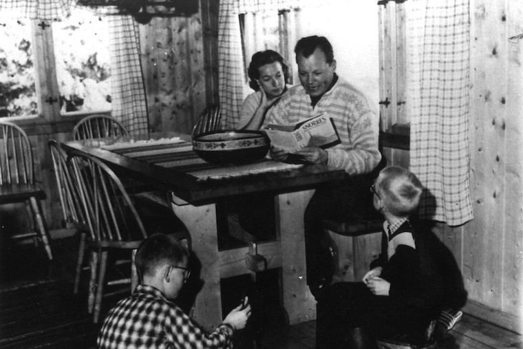 Schwarz-Weiß-Aufnahme aus dem Jahr 1960 von der Familie Brandt in einem Ferienhaus in Norwegen. Ruth und Willy sitzen am Tisch und Willy liest den Kindern Peter und Lars vor.