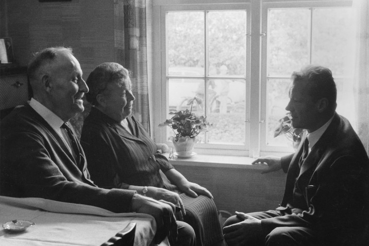 Willy Brandt sitzt bei einem Besuch in Lübeck seiner Mutter Martha Frahm und seinem Stiefvater Emil Kuhlmann gegenüber. Fotografie in Schwarz-Weiß. Öffnen, um mehr Informationen zu seiner Lübecker Familie zu erhalten.