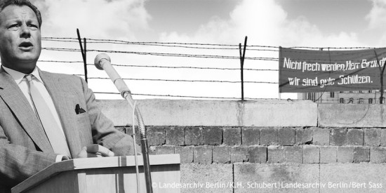 Schwarz-Weiß-Collage von Willy Brandt an einem Rednerpult und der Berliner Mauer. Hinter der Mauer steht auf einem schwarzen Transparent mit weißer Schrift: „Nicht frech werden Herr Brandt, wir sind gute Schützen.“ Öffnet den multimedialen Zeitstrahl der Jahre von 1957 bis 1966.