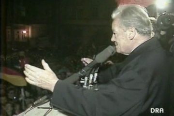 Schwarz-Weiß-Aufnahme Willy Brandts 1990, der vor mehreren Mikrofonen steht. In der rechten unteren Ecke ist das Logo DRA (Deutsches Rundfunkarchiv) zu erkennen.