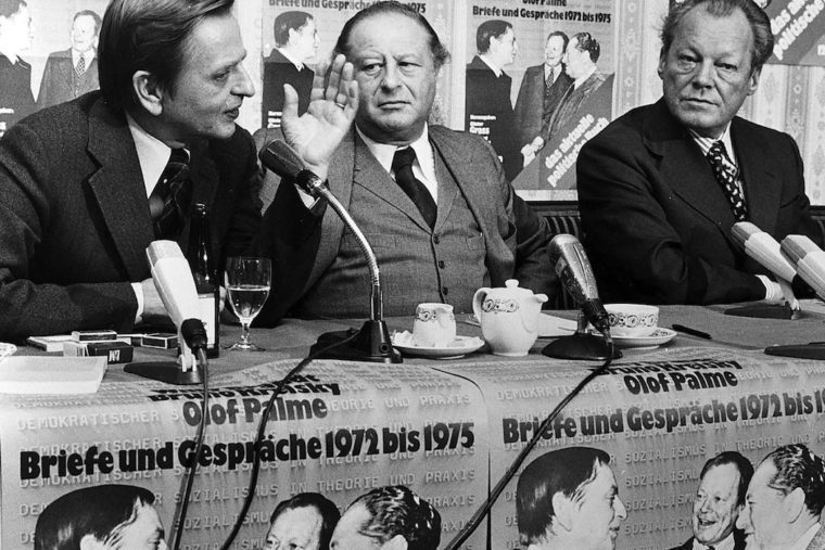 Schwarz-Weiß-Aufnahme aus dem Jahr 1975 bei der Willy Brandt neben Olof Palme und Bruno Kreisky an einem Tisch sitzt. Von den Tischen hängen Plakate mit dem Cover ihres Buches „Briefe und Gespräche 1972 bis 1975“ herunter.