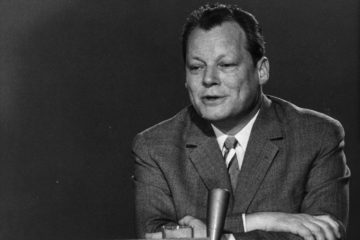 Schwarz-Weiß-Aufnahme von Willy Brandt, der im Jahr 1961 mit verschränkten Armen vor einem Mikrofon sitzt.