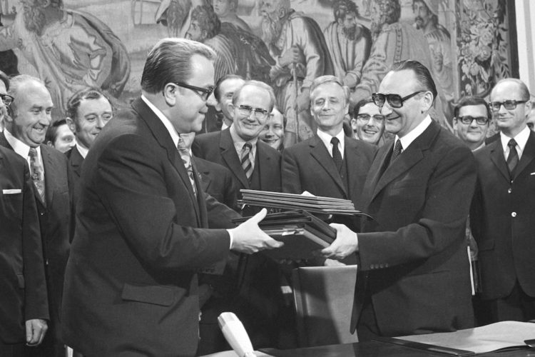 Egon Bar und Michael Kohl überreichen einander die Unterlagen des Grundlagenvertrags 1972. Im Hintergrund ist eine Reihe weiterer Politiker und Beamter zu sehen. Fotografie in Schwarz-Weiß.