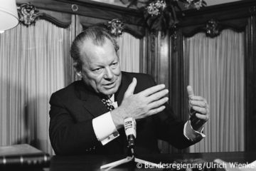 Willy Brandt sitzt hinter einem Mikrofon. Er gestikuliert mit den Händen. Fotografie in Schwarz-Weiß. Öffnet Originaltöne von Reden und Interviews.