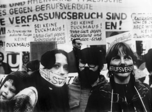 Schwarz-Weiß-Aufnahme einer Demonstration im Jahr 1977, die sich gegen Berufsverbote richtet. Zu sehen sind im Vordergrund zwei Frauen, die sich den Mund verbunden haben, darauf steht „Berufsverbot“. Im Hintergrund sind Plakate zu erkennen auf denen unter anderem steht: „Keine Duckmäuse“, „Sei keine Duckmaus“.